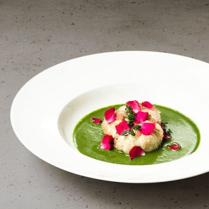 Ein Teller mit einem in Rosenwasser gedämpften Igestachelbart-Pilz (Hericium erinaceus) – auch Löwenmähne, Affenkopfpilz oder Pom-Pom blanc genannt –, umgeben von Moretum – einer Art Pesto aus dem antiken Rom. Dekoriert mit Rosengel, frischen Rosenblüten und einem Zweig Weinraute (Ruta graveolens).