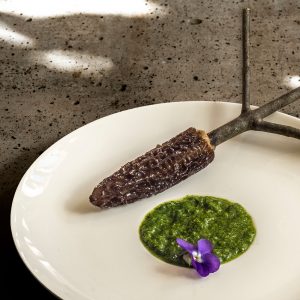 Ein weißer Teller mit einer gefüllten Morchel "Bengali Style" auf einen Ast aufgespießt und einem Klecks Koriander-Minz-Chutney und einer Duftveilchenblüte.